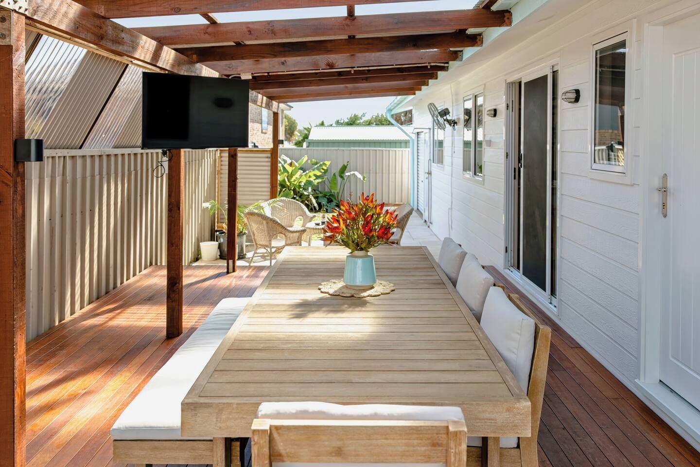 An outdoor patio with a wooden table and chairs, a TV mounted on a wooden beam, and a white fence in the background.