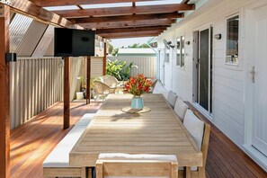 An outdoor patio with a wooden table and chairs, a TV mounted on a wooden beam, and a white fence in the background. - Casa Aqua: Tropical/Coastal Escape & McLaren Vale (Christies Beach)