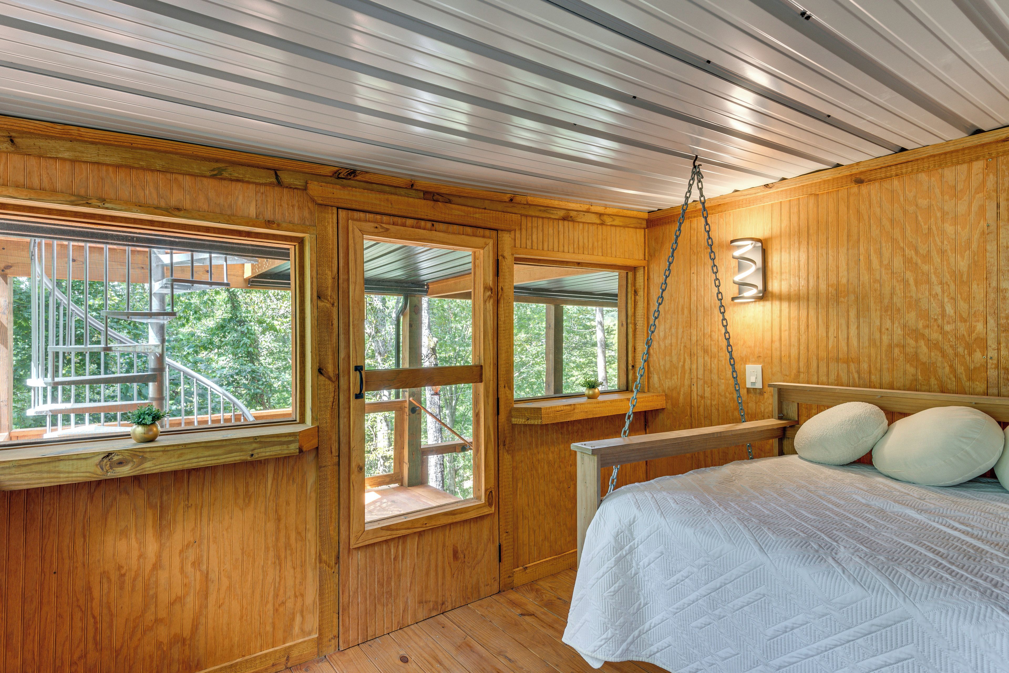 A wooden cabin bedroom with a bed, two windows, a small balcony, and a hanging light fixture.