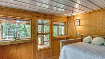 A wooden cabin bedroom with a bed, two windows, a small balcony, and a hanging light fixture.