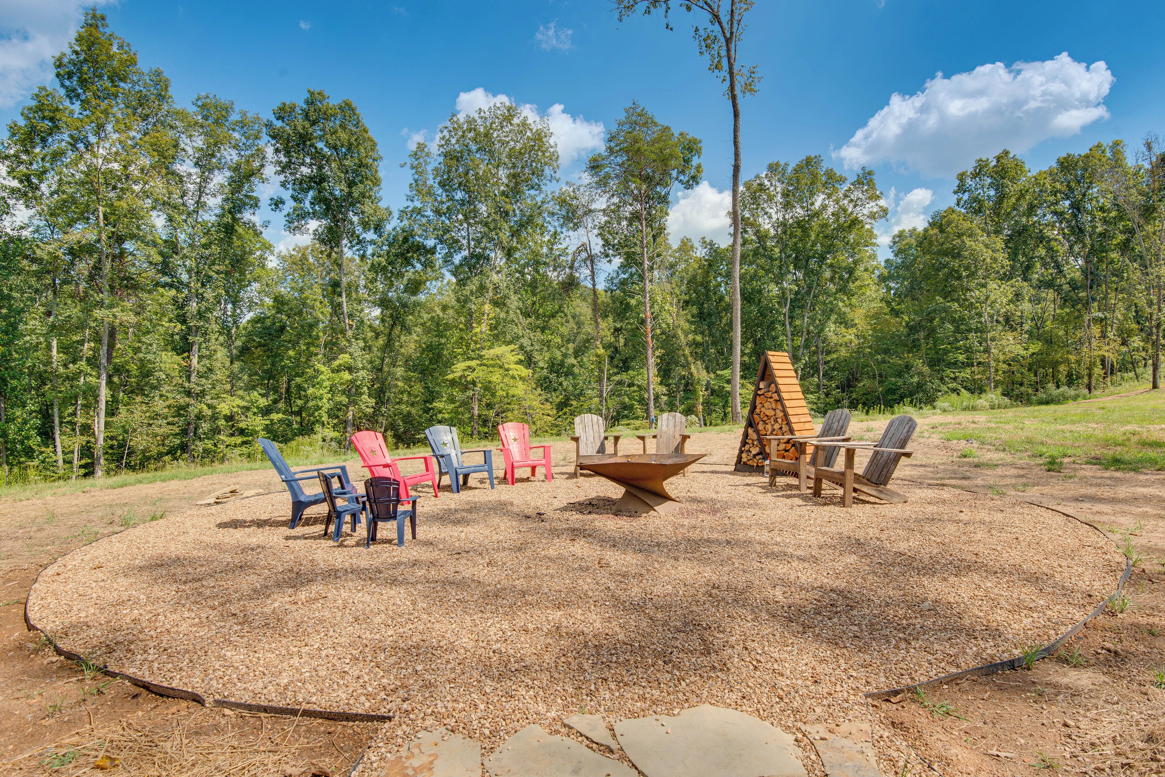 A circular outdoor seating area with Adirondack chairs and a fire pit, surrounded by a forested landscape.