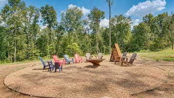 A circular outdoor seating area with Adirondack chairs and a fire pit, surrounded by a forested landscape.