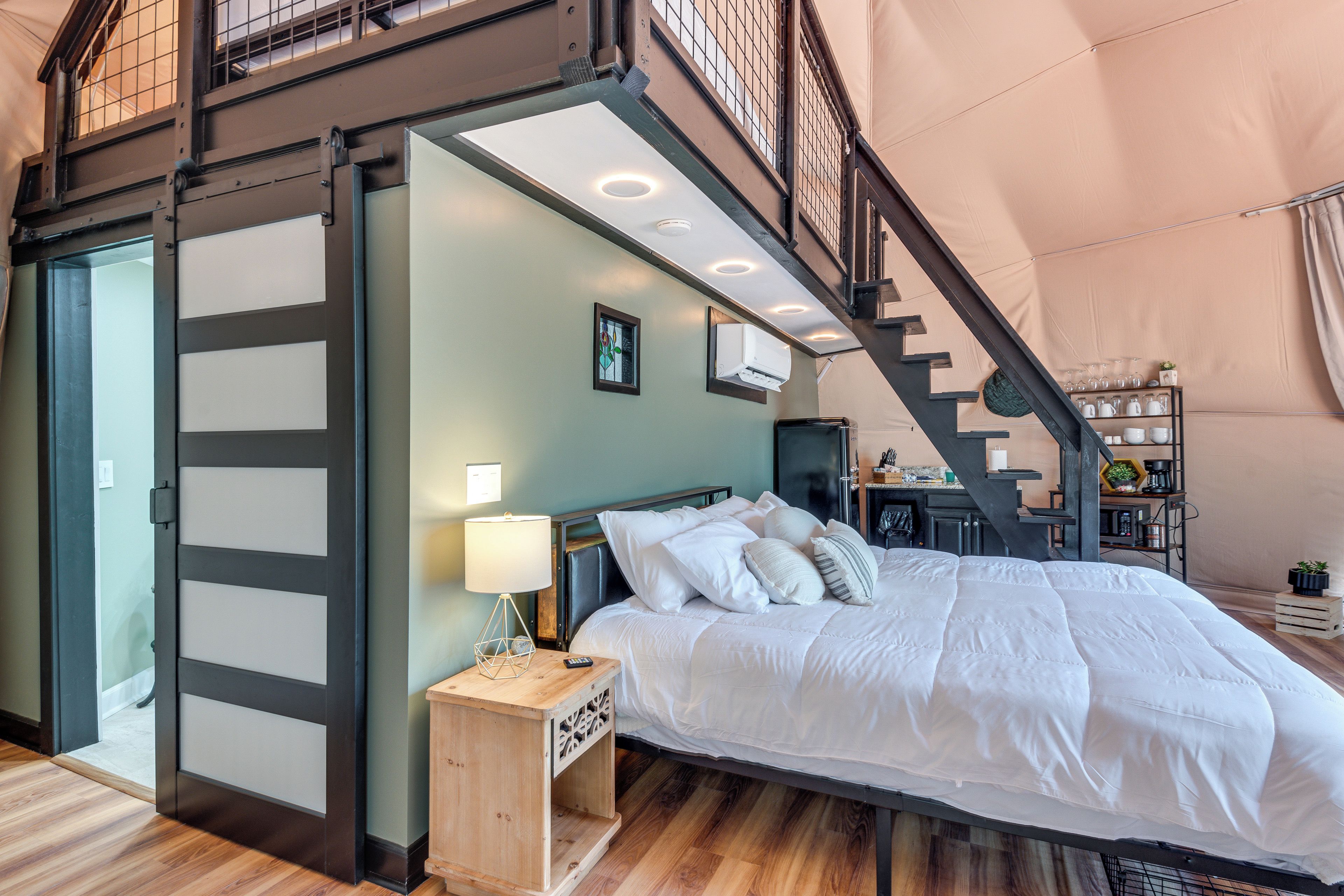 Quarto com cama, mesa de cabeceira, abajur e escada para um andar superior.