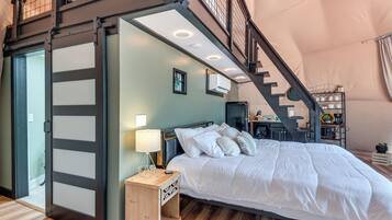 Quarto com cama, mesa de cabeceira, abajur e escada para um andar superior.