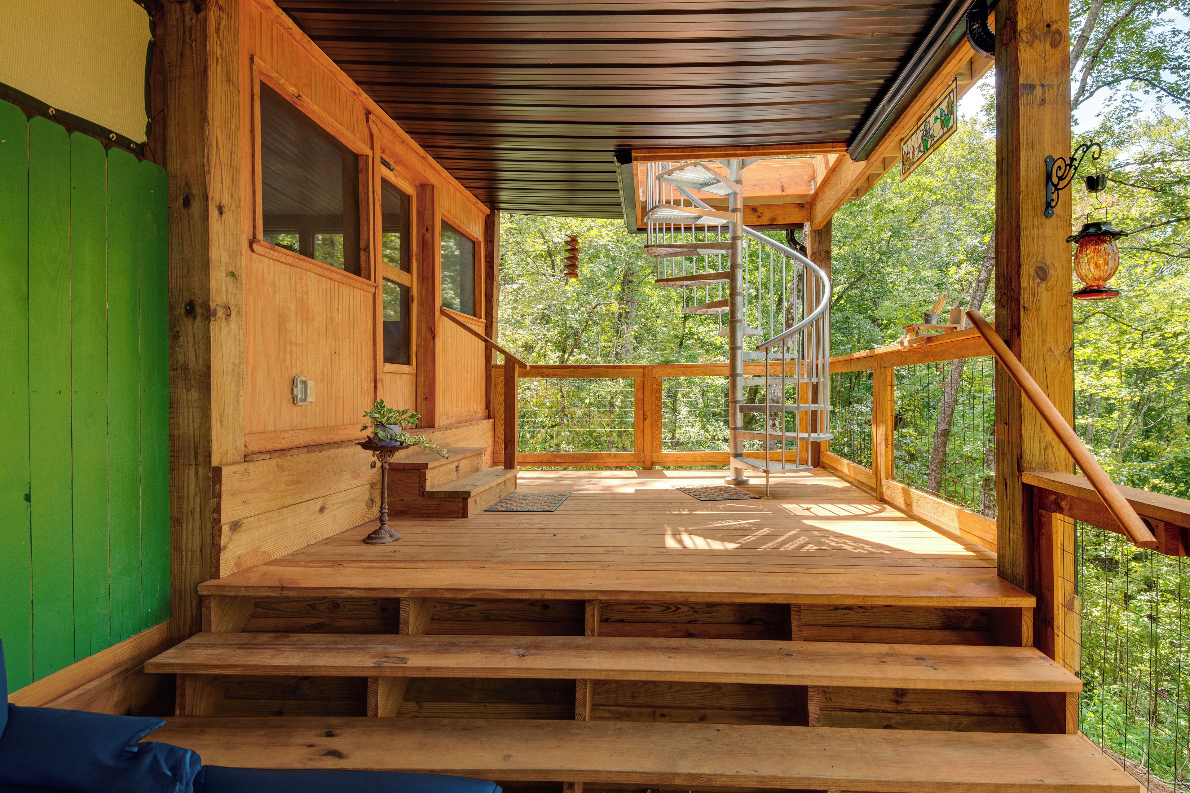 Una escalera de madera lleva a una terraza cubierta con una escalera en espiral, rodeada por una barandilla de madera y con vistas a un área boscosa.