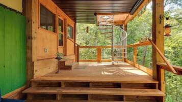 Una escalera de madera lleva a una terraza cubierta con una escalera en espiral, rodeada por una barandilla de madera y con vistas a un área boscosa.