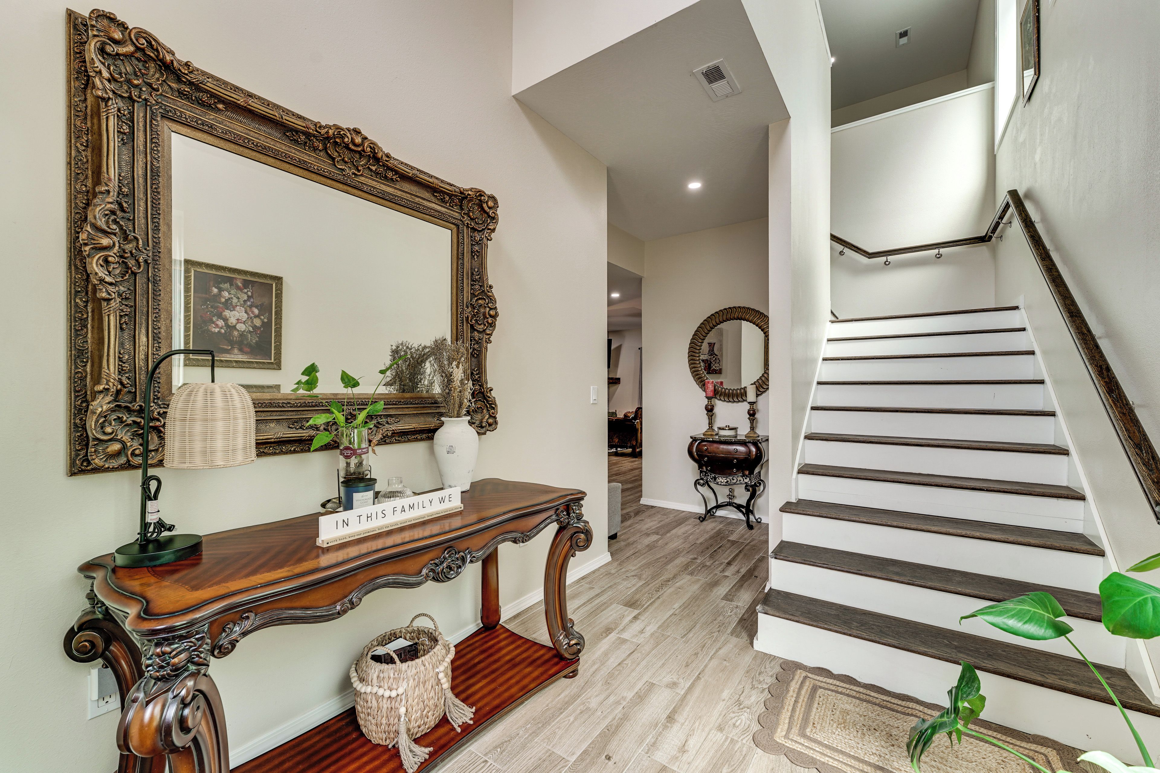 A hallway with a wooden console table, a mirror, a vase with plants, and a staircase.