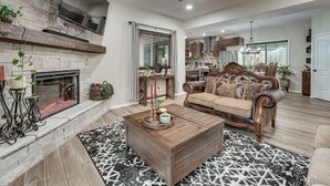 A living room with a fireplace, a sofa, a coffee table, and a kitchen in the background. - Patio, Porch + Fire Pit! Family Home By Branson (Rockaway Beach)