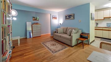 A living room with a sofa, a coffee table, and a bookshelf. There is a kitchen area with cabinets and a countertop.