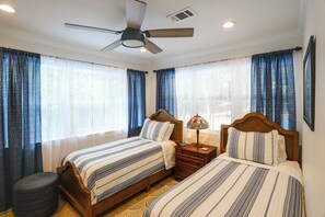 Quarto com duas camas, um ventilador de teto, mesa de cabeceira com abajur e janela com cortinas.