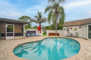 Piscina com borda azulejada, área de estar coberta com sofá e almofadas, e um pequeno deck com cadeiras.