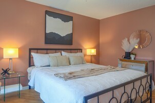 Una cama bien hecha con edredón blanco y varios almohadones, una mesita de noche con lámpara, un cuadro abstracto en la pared, y un espejo redondo con marco decorativo.