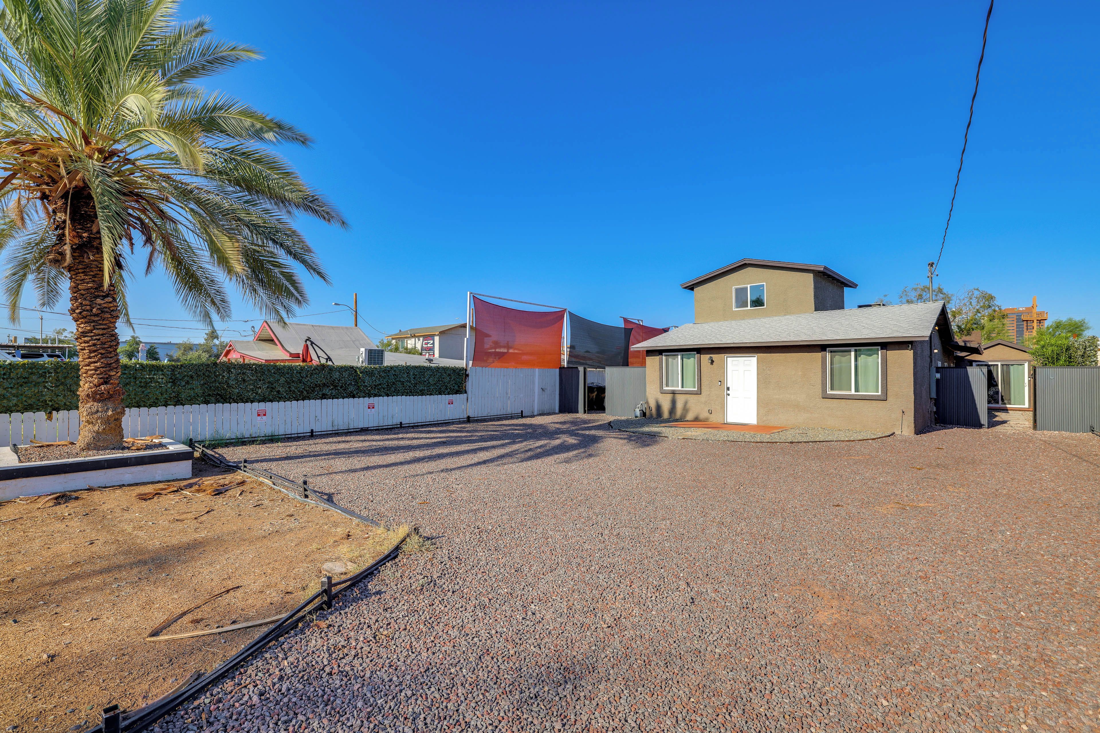 Un barrio residencial con camino de grava, una palmera, patio cercado y una casa de dos pisos con techo rojo.