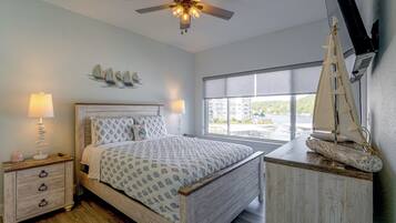 A bedroom with a bed, bedside lamps, a ceiling fan, a sailboat decoration, and a window with a view.