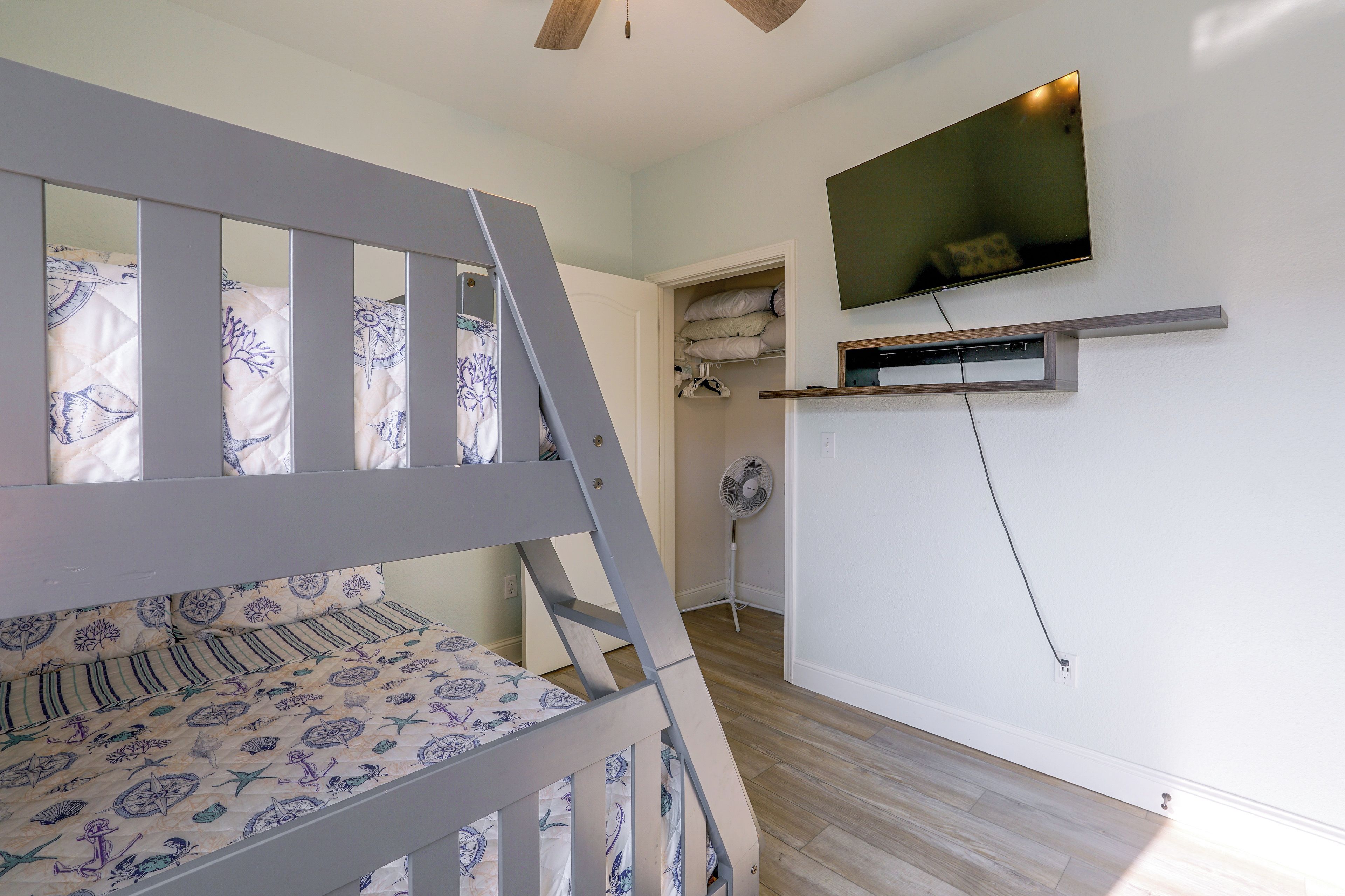 A bedroom with a bunk bed, a flat-screen TV mounted on the wall, and a standing fan.