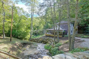 A rustic cabin surrounded by a dense forest, with a stone wall and a small stream nearby.