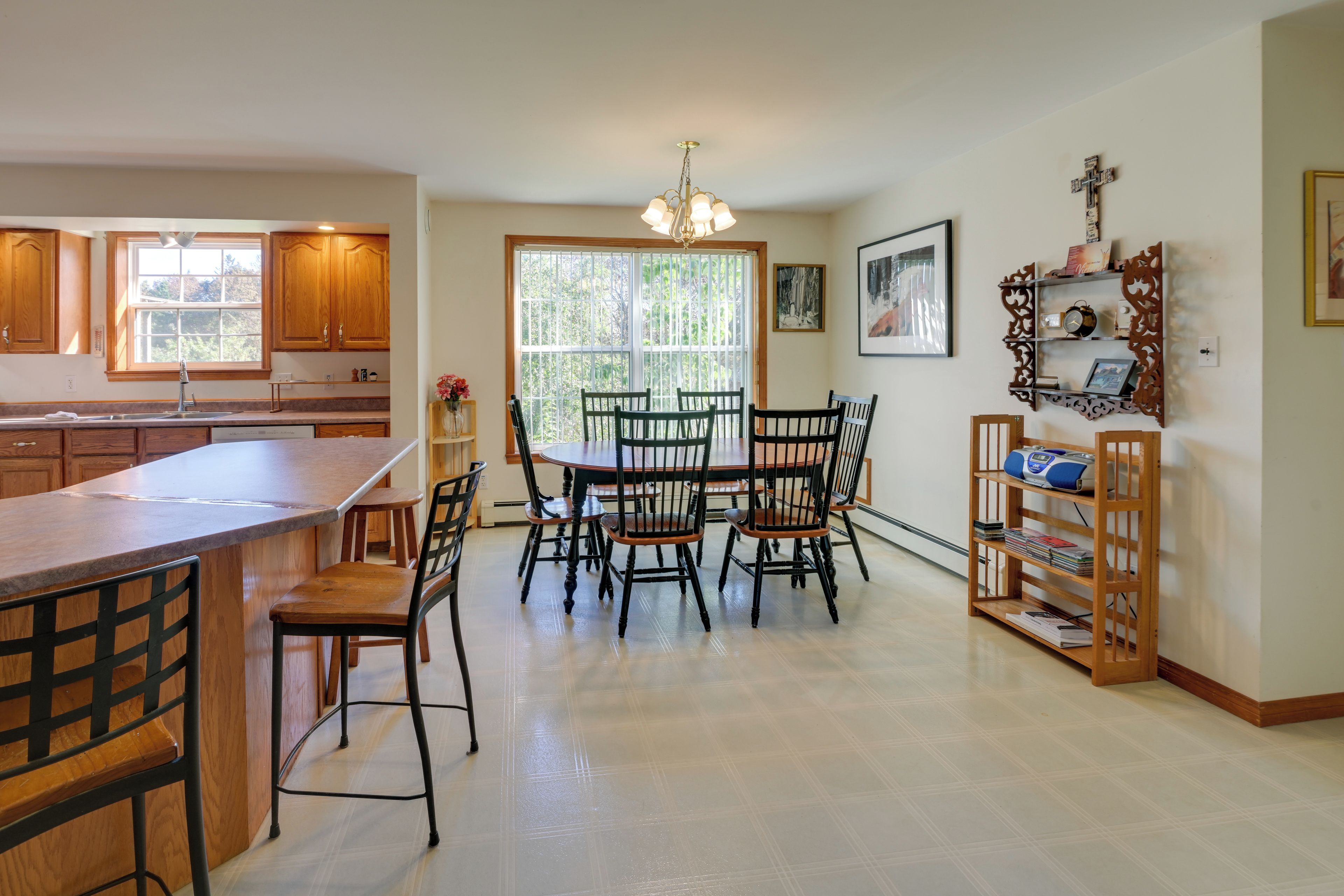 A kitchen with wooden cabinets, a dining table with chairs, a built-in bookshelf, and a window with a view of greenery.
