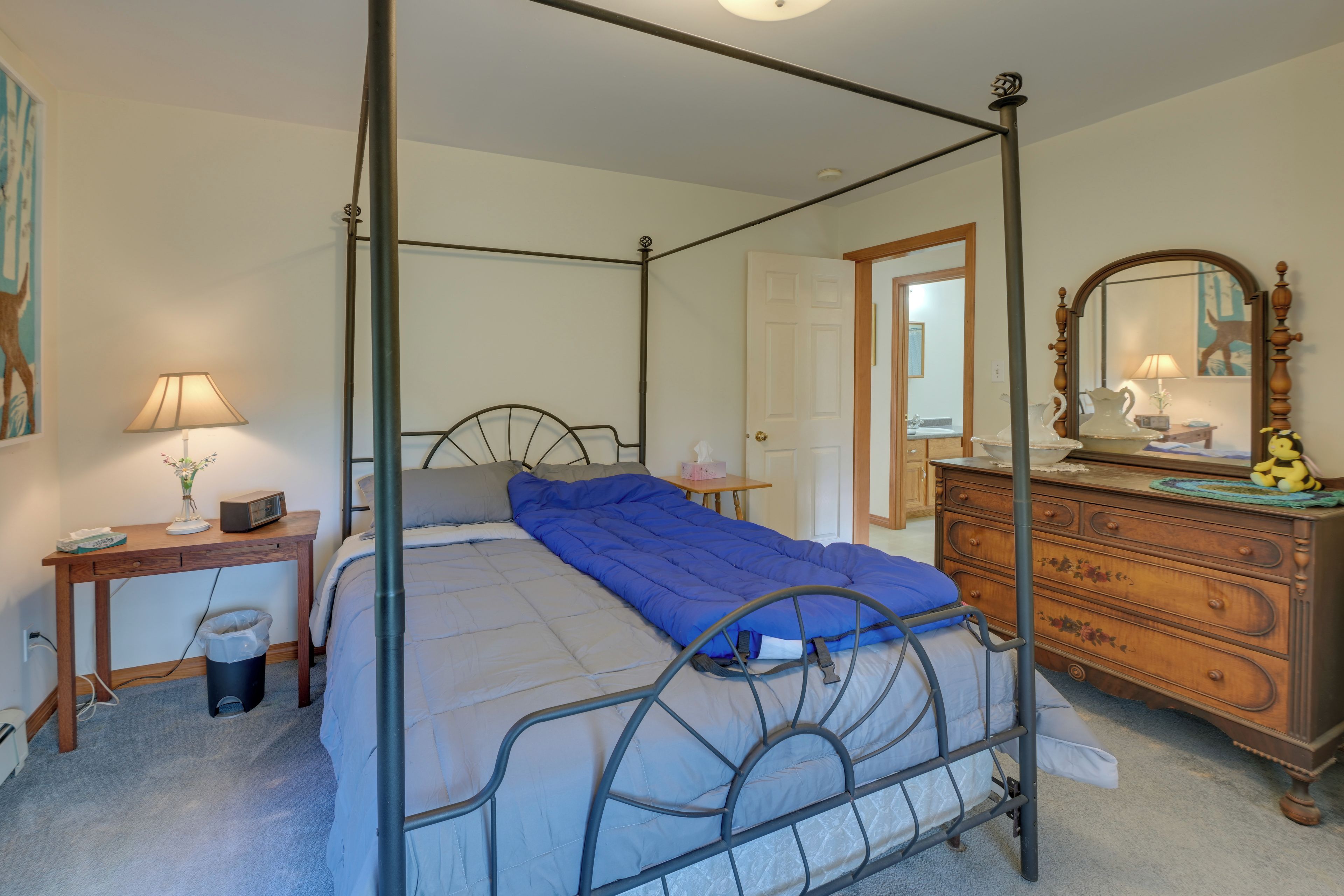 A four-poster bed with a blue blanket, a wooden dresser with a mirror, a bedside table with a lamp, and a trash bin.