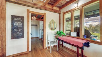 A room with wooden beams, a table with a red tablecloth, a chair, a mirror, and a window with a view of trees.