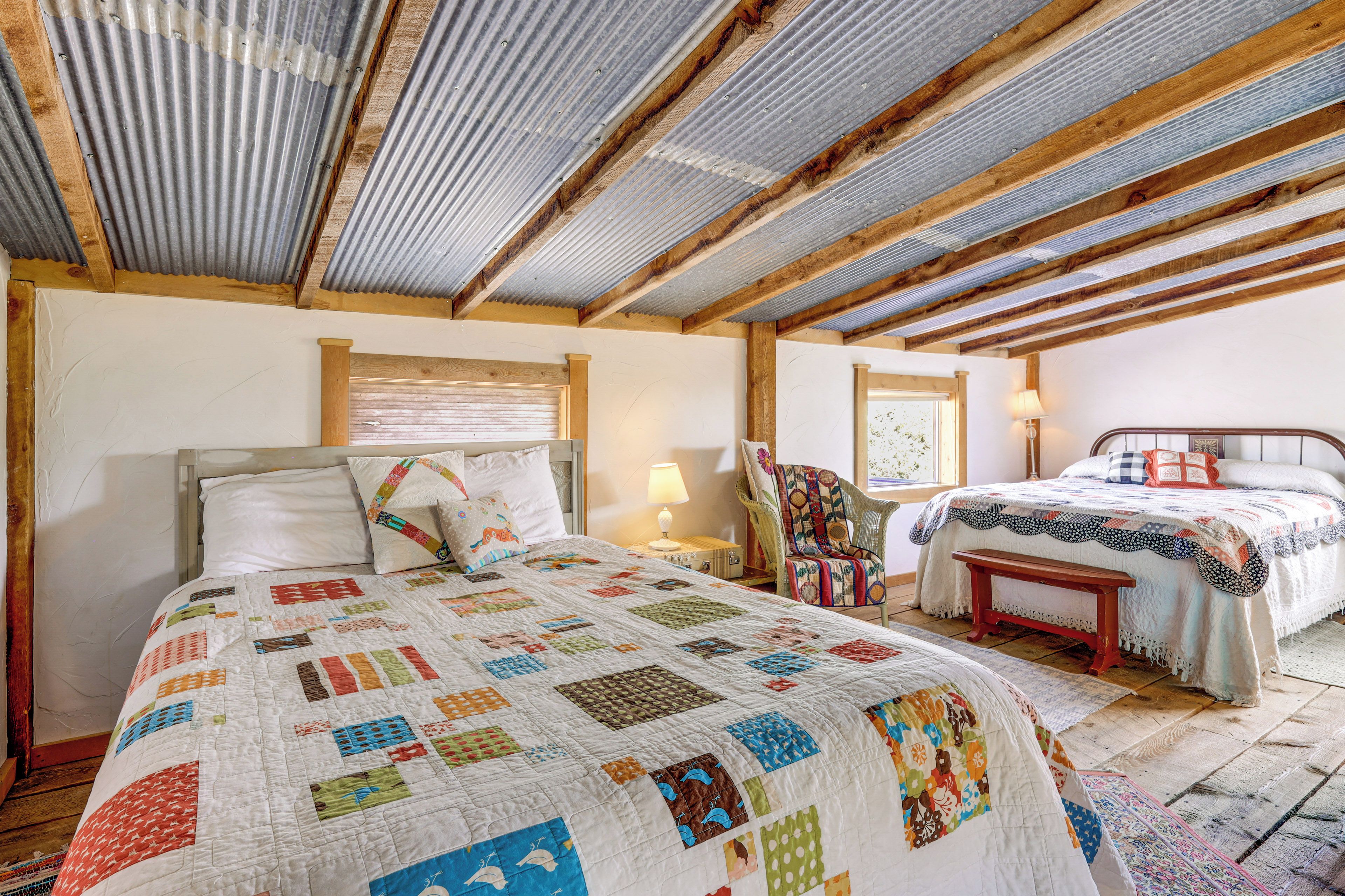 A bedroom with a wooden ceiling, a large bed with a colorful quilt, a smaller bed, a chair, and a bedside table.