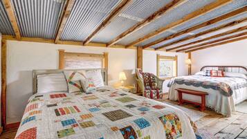 A bedroom with a wooden ceiling, a large bed with a colorful quilt, a smaller bed, a chair, and a bedside table.