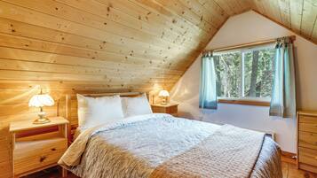 Un dormitorio con techo de madera, una cama con ropa de cama blanca, dos lámparas de noche, una ventana con cortinas y una mesita de noche de madera.