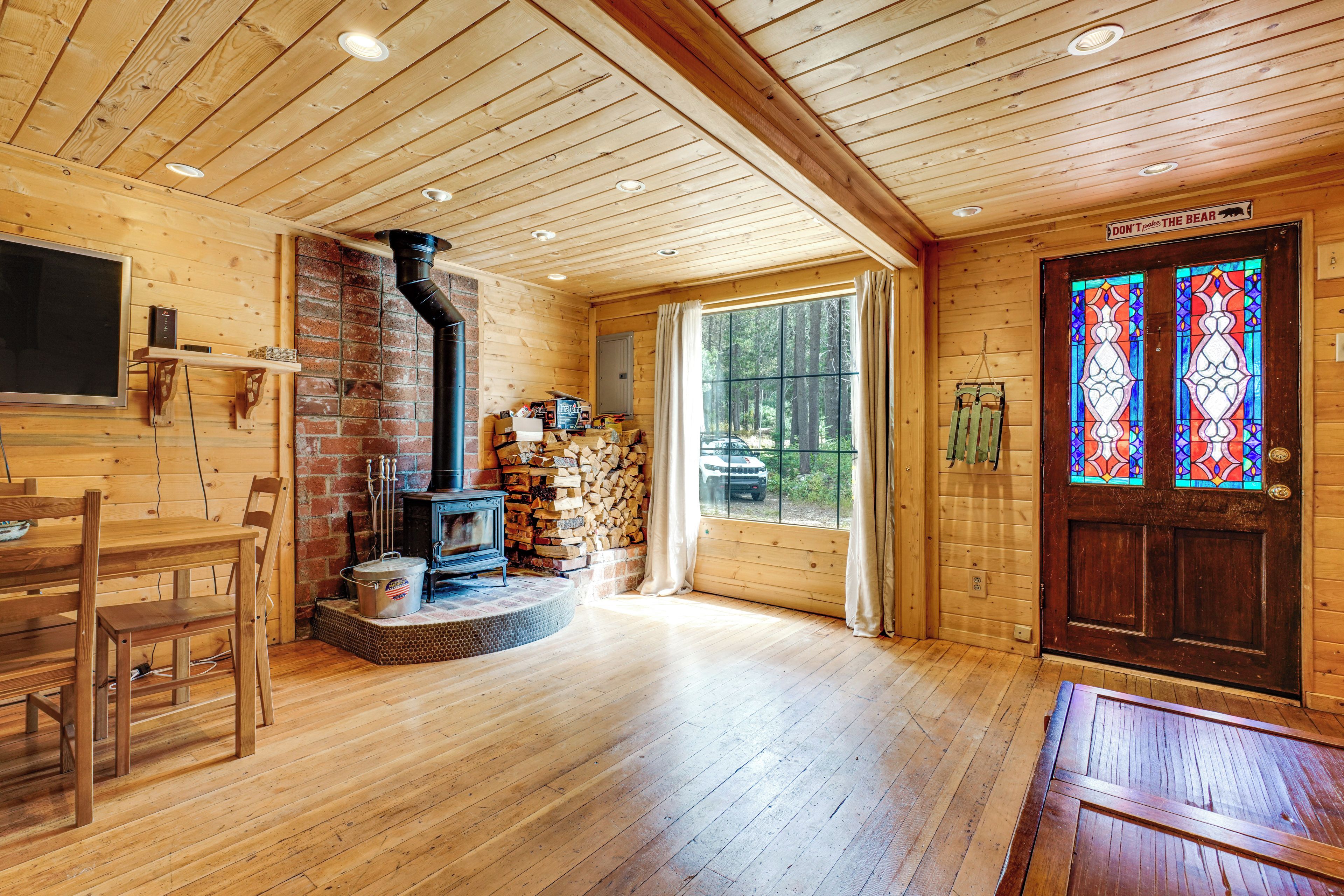 A cozy cabin interior with a fireplace, wooden walls, a dining area, and a large window with a view of the forest.