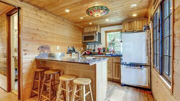 Interior de una cabaña de madera con cocina, que incluye refrigerador, estufa y taburetes de bar.