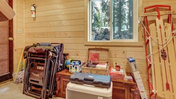 A room with wooden walls, a window with a view of trees, a fire extinguisher, and various items on a table including a basket, a cooler, and a badminton racket.