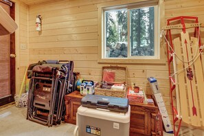 A room with wooden walls, a window with a view of trees, a fire extinguisher, and various items on a table including a basket, a cooler, and a badminton racket. - Cozy Mountain Cabin Skiing Hiking Biking Swimming (Truckee)