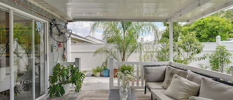 A covered outdoor seating area with a sofa and a table, surrounded by potted plants and a view of the neighborhood.
