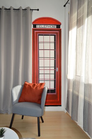 A room with a telephone booth-themed door, a grey armchair with an orange cushion, and sheer curtains.