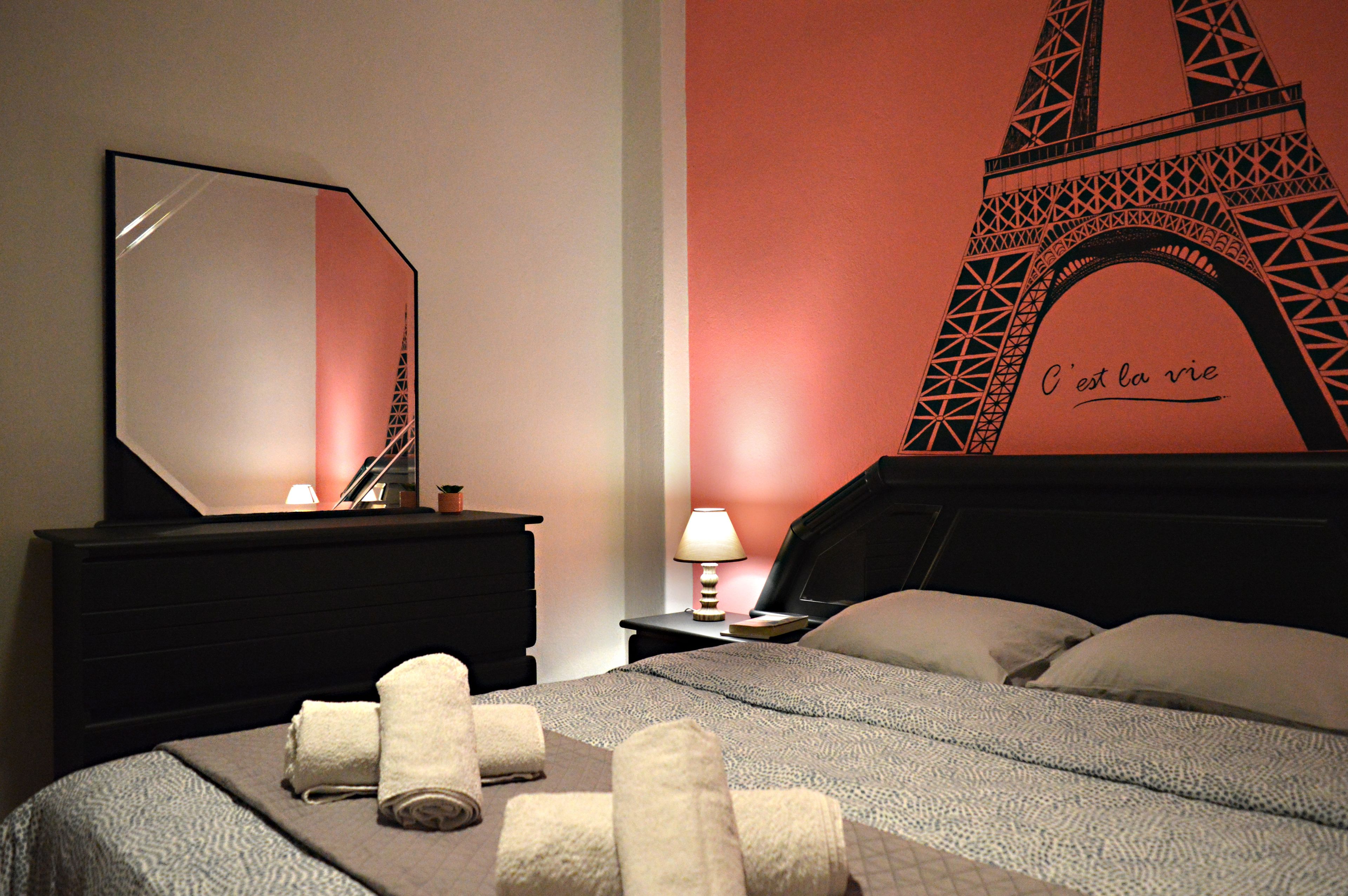 A hotel room with a bed, a dresser, a mirror, a lamp, and a wall with a large Eiffel Tower mural.