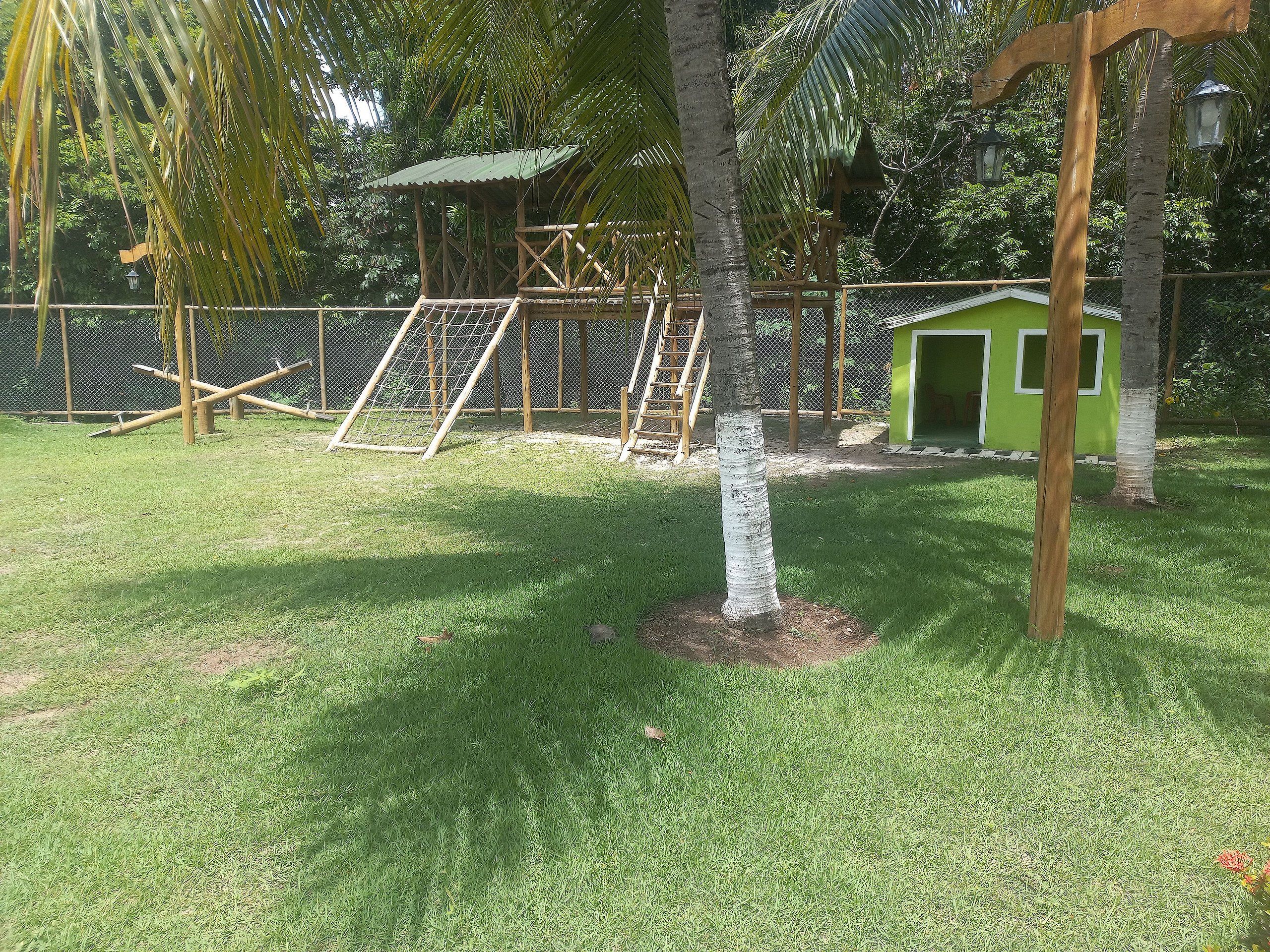 Uma área externa cercada com uma pequena construção verde, um parquinho e uma árvore.