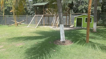 Uma área externa cercada com uma pequena construção verde, um parquinho e uma árvore.