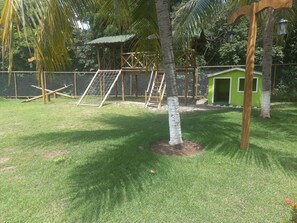 Un espacio exterior cercado con una pequeña estructura verde, un parque infantil y un árbol.