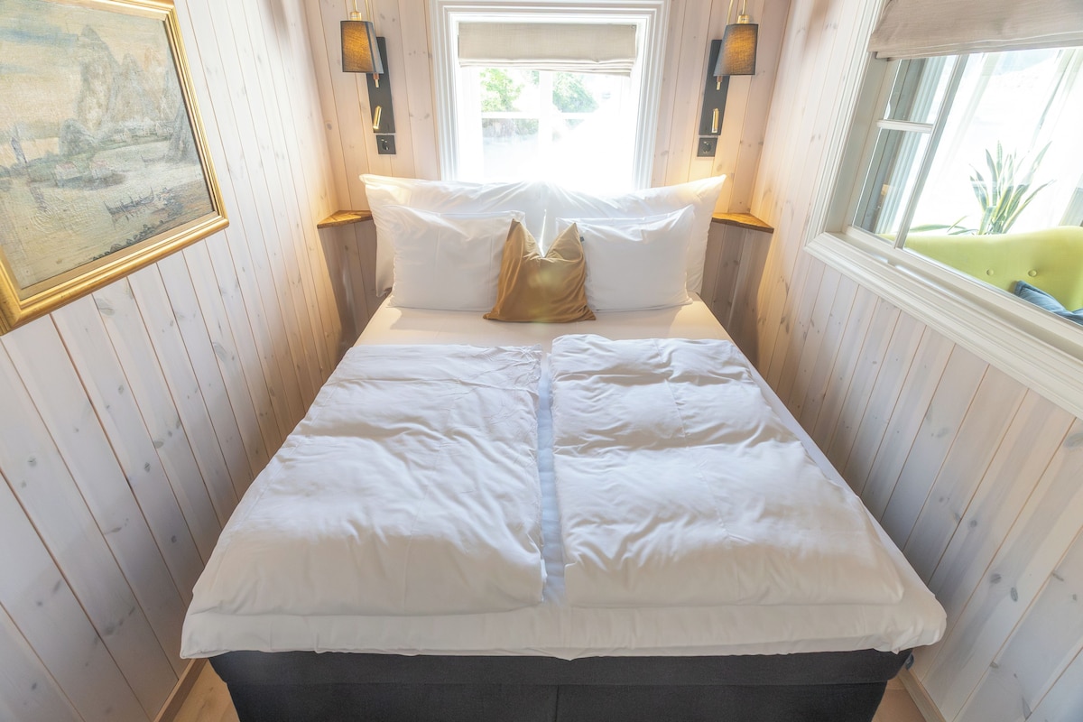 Cama com roupa de cama branca, duas almofadas e uma almofada decorativa. Há luminárias fixadas na parede, de cada lado de uma janela.