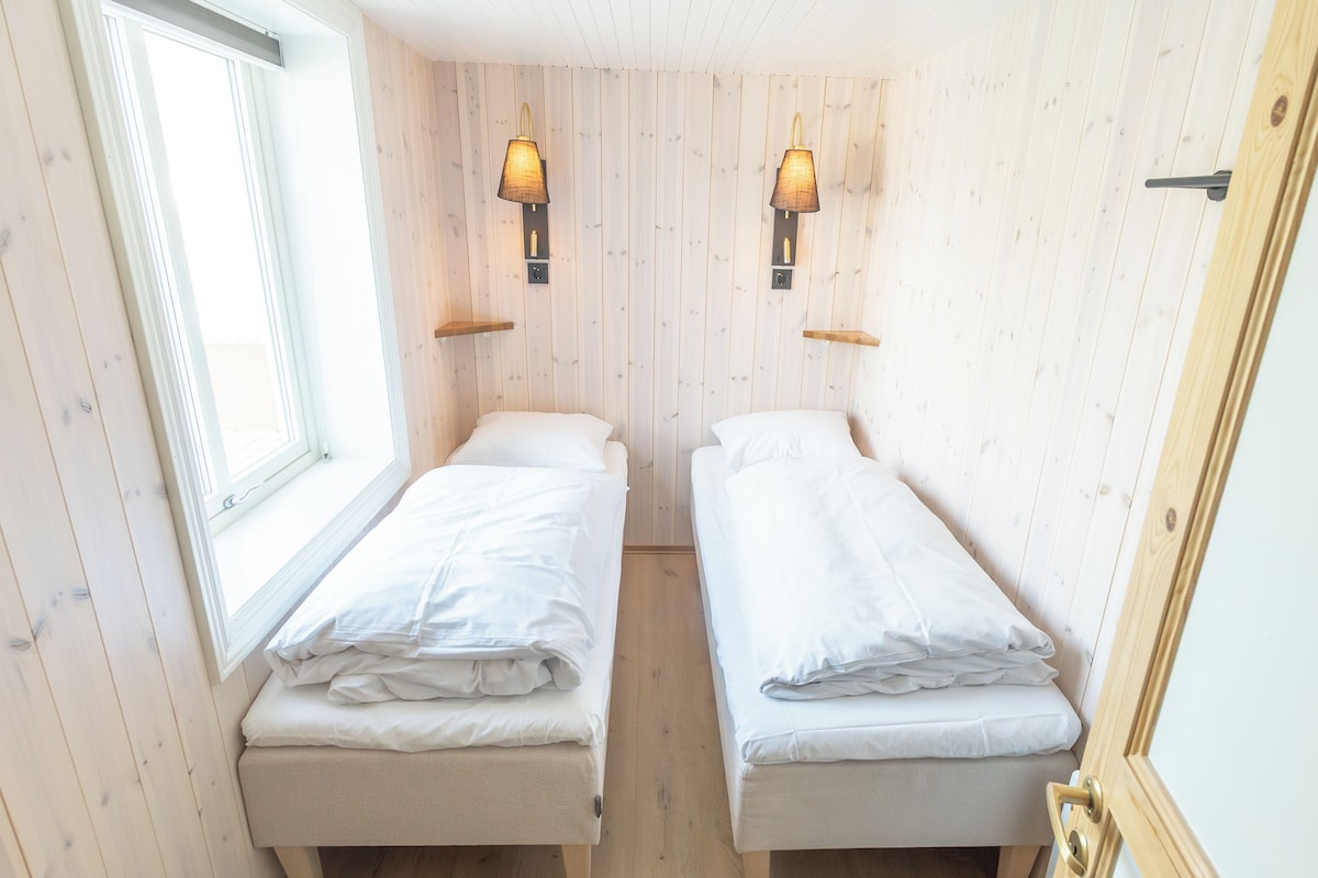 Acomodação pequena, com um quarto e duas camas, janela e iluminação fixa na parede.
