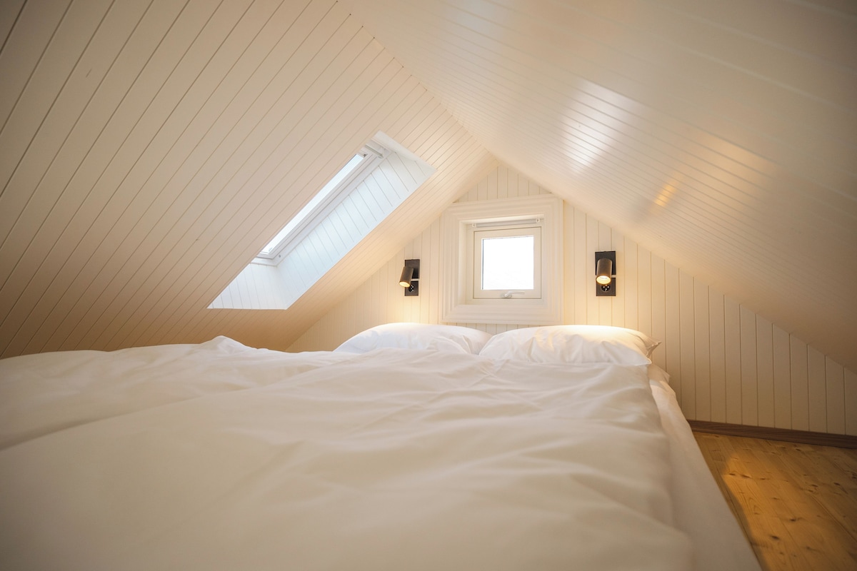 Quarto com teto inclinado, claraboia, cama e duas luminárias.