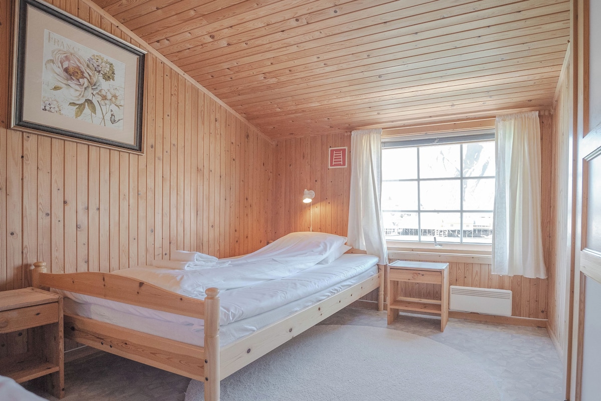Quarto de madeira com uma cama, uma escrivaninha e uma janela com cortinas.