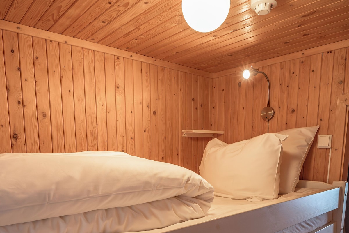 Uma cama de beliche com roupa de cama branca num quarto de madeira com teto inclinado e luminária fixada na parede.