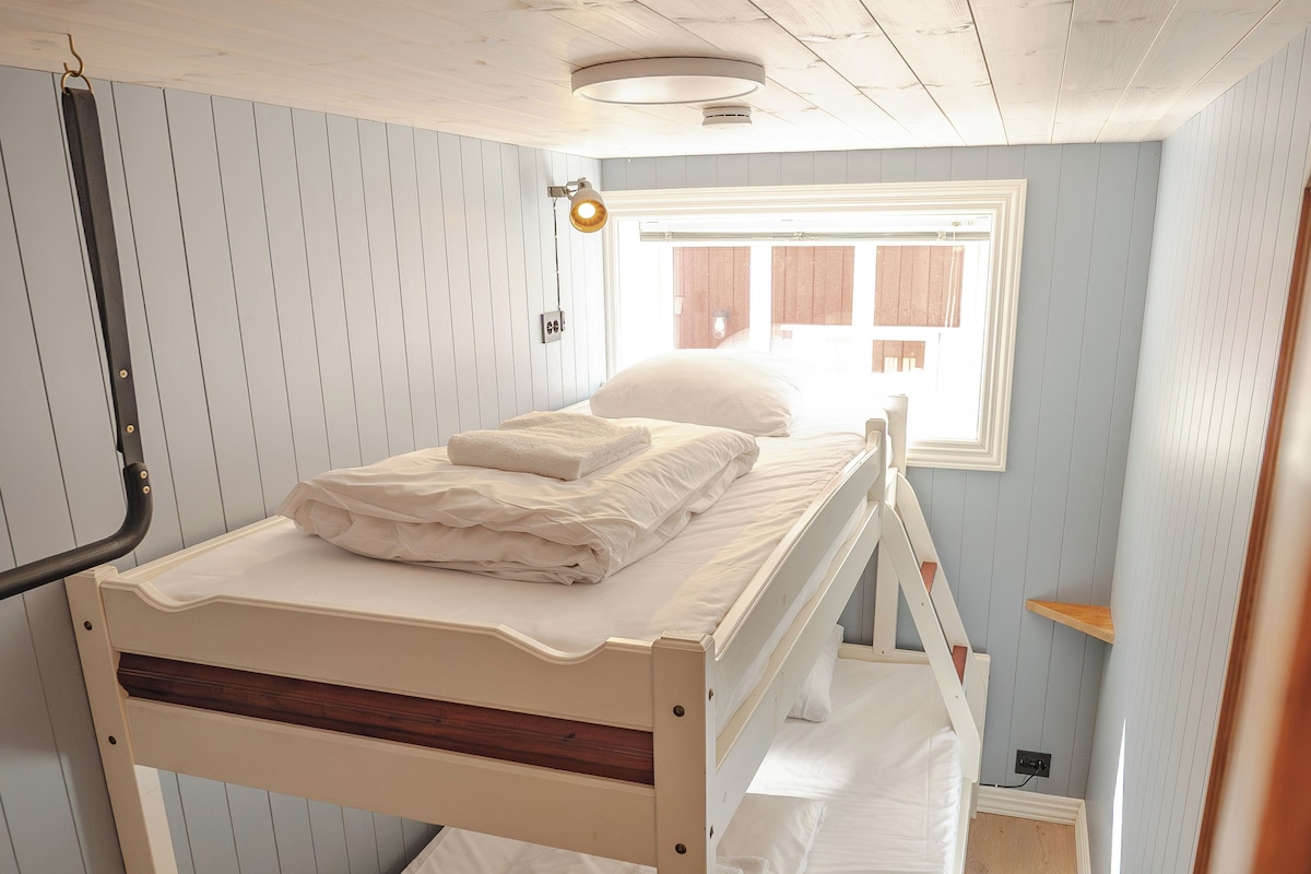 Uma cama beliche com roupa de cama branca, um banco de madeira e uma janela com persianas.