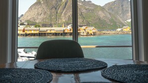 A view from a window with a table and chairs, overlooking a body of water with buildings and mountains in the background. - Rostad Retro Rorbuer (Nordland)