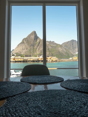 Vista da janela com uma mesa e cadeiras, sobre um corpo d'água com edifícios e montanhas ao fundo.