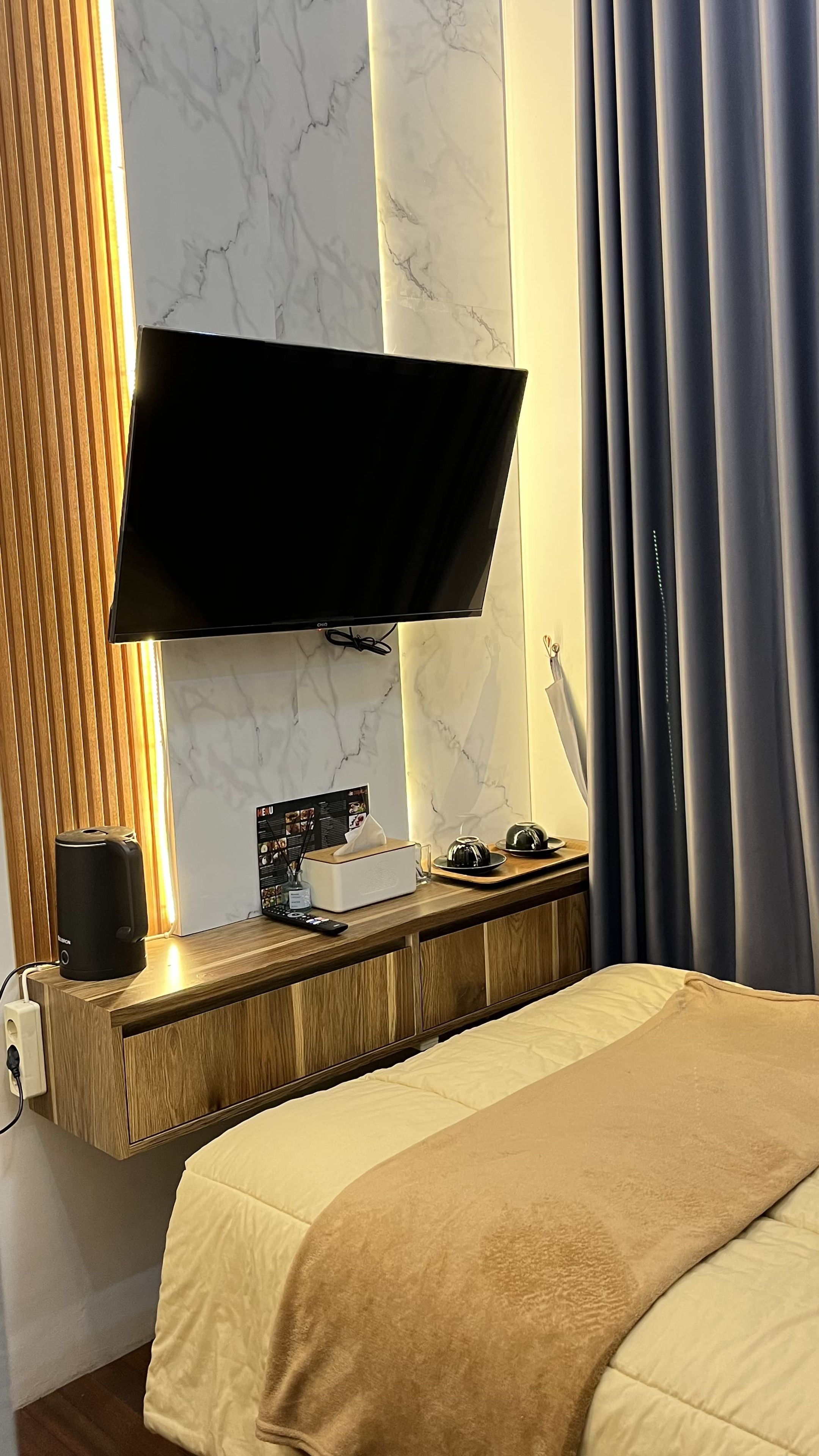 Ein Hotelzimmer mit einem an der Wand montierten Flachbildfernseher, einem Nachttisch aus Holz inklusive Fernbedienung, einer Taschentuchbox und einem kleinen dekorativen Gegenstand.