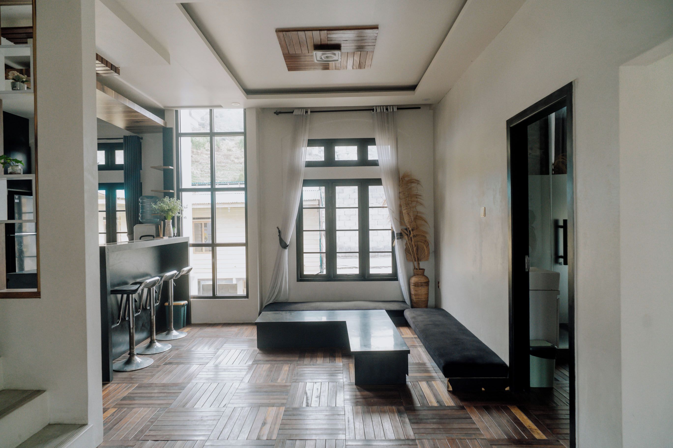 A modern interior with a wooden herringbone floor, a black sofa, a dining area with bar stools, and large windows with curtains.