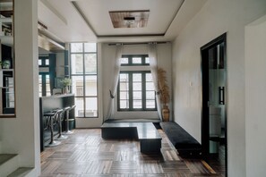 Un interior moderno con suelo de parqué, un sofá negro, una zona de comedor con taburetes altos y ventanas grandes con cortinas.
