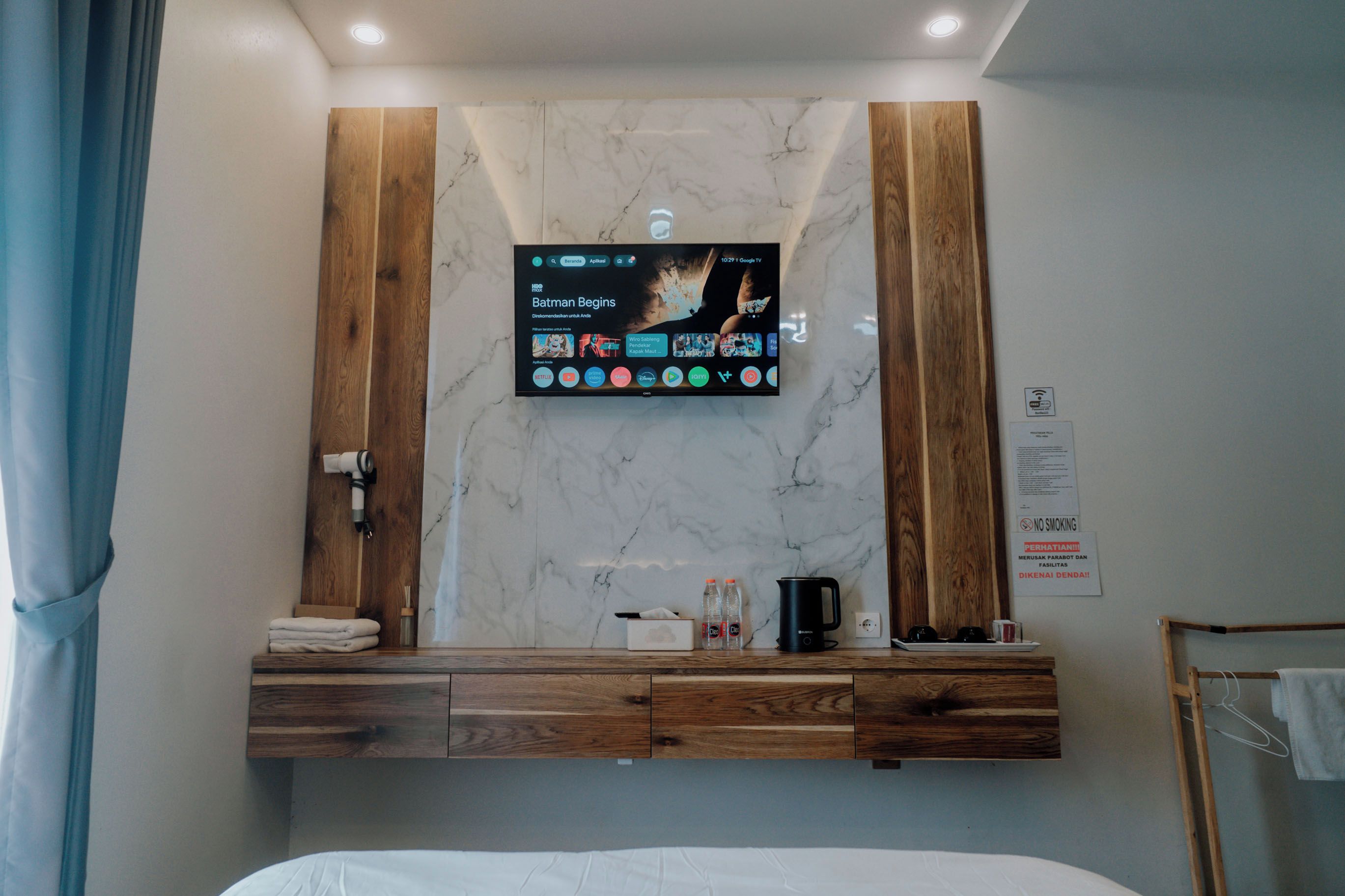 Een hotelkamer met een grote flatscreen-televisie bevestigd aan een marmeren muur, houten paneelwanden, een nachtkastje met een waterkoker en een bed.