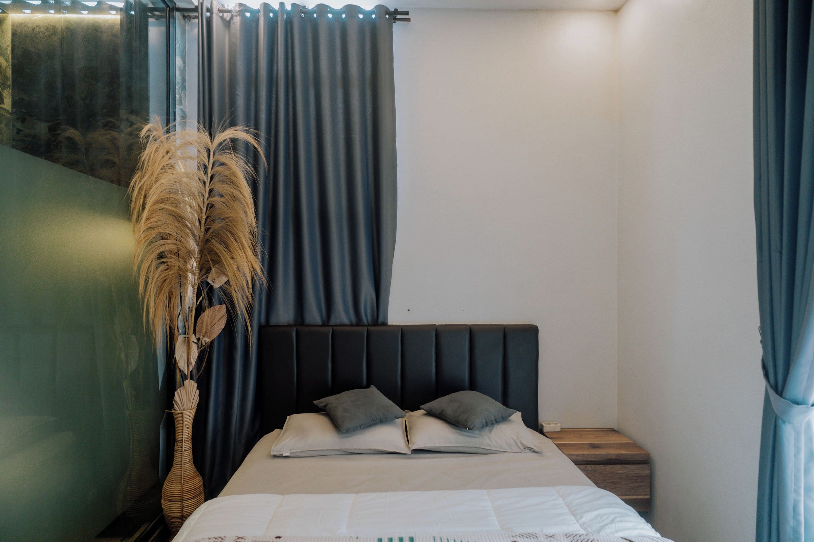 A bedroom with a bed, pillows, a nightstand, a vase with dried plants, and blue curtains.