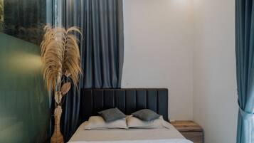 A bedroom with a bed, pillows, a nightstand, a vase with dried plants, and blue curtains.